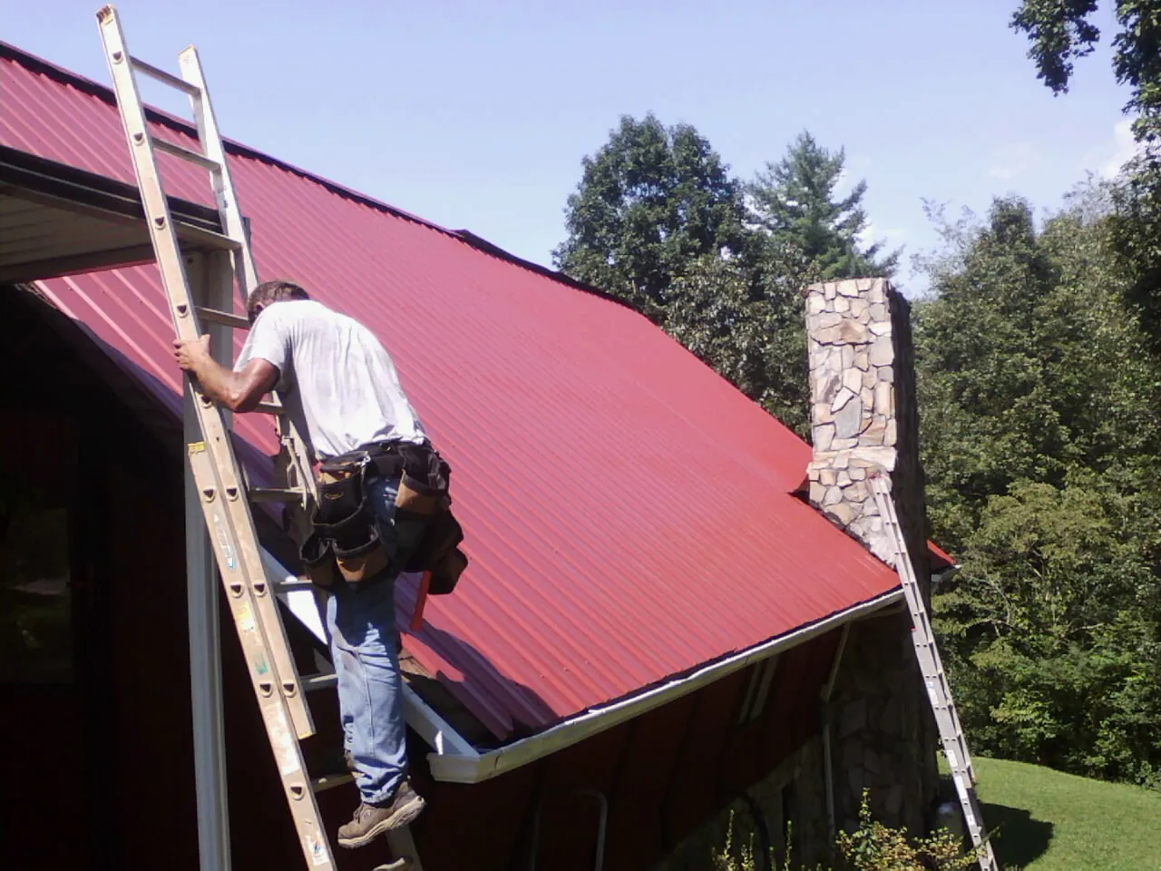 Roofer on a ladder beside a red metal roof and stone chimney — Soffit & Fascia Repair by Roofing Brothers in Urbana